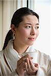 Taiwanese mid adult woman in bathrobe drinking coffee and looking out window.