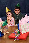 Hispanic brother and sister wearing sunglasses and party hats sitting with presents having a birthday party.