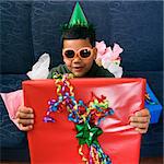 Hispanic boy wearing party hat and sunglasses holding large birthday present smiling and looking at viewer.