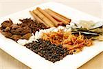 An assortment of fragrant, richly flavored spices - bay leaves, mace, peppercorns, black pepper, silver cardamom pods, cloves and cinnamon. Focus on black peppers.