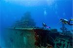 Two divers are swimming towards the superstructure of a shipwreck. Tangled wreckage in the foreground; the vessel recedes into the ocean blue in the background.  Space for copy above.