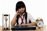 Young Chinese girl wearing school uniform sitting at desk with hand on chin  with a pouting face expression with an alarm clock and hour glass