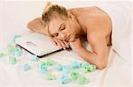 Blond naked woman lying down awaiting spa treatment, surrounded by aromatherapy items and with her head resting on weight scales