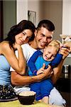 Caucasian family with toddler son in kitchen at breakfast smiling at viewer.
