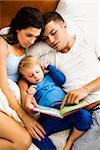 Caucasian parents and toddler son reading book in bed.