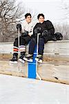 Two boys in ice hockey uniforms sitting on ice rink sidelines looking and smiling.