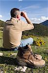 Man sitting in a meadow in the mountains looking through binoculars
