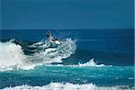 young man swings his surf board around as he tops a large wave.  Blue skies and blue water.