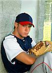 young teen sits on the ground and leans back against dugout wall.  He is unsmiling and is holding a glove in his hands.  Uniform is in navy, white and red.