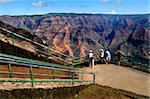 Grand Canyon of Kauai, Hawaii has deep gorges and ridges in hues of pink and orange.  This photo has tourists enjoying the view.  Two men and two women look over rail.  Two are aiming cameras.
