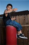 Young asian boy sitting on top of a punching bag outside beside a tall wooden fence smiling wearing jeans and black tshirt