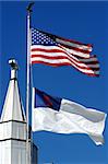 A cross tops the steeple of a church.  Two flags fly in front of the cross.  One is the American Flag and the other is the Christian Flag.  Blue sky surrounds both.