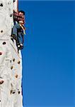 Young asian boy outside climbing on a rock climbing wall with clear blue sky in background