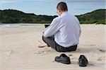 Businessman working with his laptop while sitting barefoot on the beach.