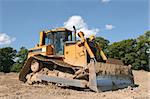 Yellow bulldozer standing idle on rough earth with trees and a blue sky to the rear.