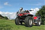 Four wheel drive red and black quad bike standing idle on the grass, with trees and a blue sky with clouds to the rear.