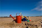 Red watering can, plastic blue bucket and other beach toys in the sandy seashore