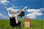 Business concept shot of a beautiful young woman sitting at a desk using a computer in a green field raising her arms into a bright blue sky with fluffy white clouds. Shot on location with copyspace at the top of the image.