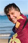 A handsome smiling forties man is sat on a beach with a towel wrapped around him to keep warm.