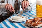 Bavarian man dressed in traditional leather trousers (lederhosen) is eating a veil sausage (Weisswurst) and beside him is a full beer stein (Maß) and pretzel.