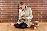 women in a trenchcoat sitting next to a pile of books and reads one