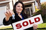 Happy Attractive Hispanic Woman Holding Keys and Sold Sign In Front of House.