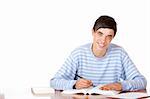 Young student is sitting on desk with open book and learns for his exams. He looks smiling into camera. Isolated on white.