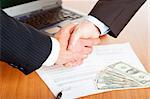 Handshake of businessmen with laptop, us dollar and contract in background.