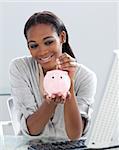 Assertive Afro-american businesswoman saving money in a piggybank at her desk