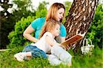 young mother sitting in grass under tree and reading book to her small daughter who is lying on knees of her mother and smiling
