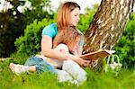 young mother sitting in grass under tree and reading book to her small daughter who is lying on knees of her mother
