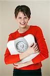 Attractive young woman holds a bathroom scale to her chest while smiling at the camera. Vertical shot.