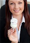 Happy businesswoman holding a light bulb smiling at the camera sitting in her office