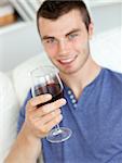 Charismatic young man holding a glass of wine sitting on a sofa in the living-room