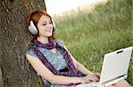 Young smiling fashion girl with notebook and headphones sitting near tree.
