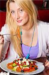 A young woman sitting in a restaurant. She is looking at the camera and smiling as she is eating salad. Vertical shot.
