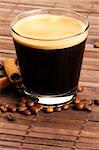 espresso in a short glass with coffee beans and cinnamon sticks from diagonal top on wooden background