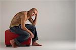 A young, muscular Brazilian man in a studio shot, on a red seat, on a gray background.