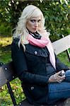 young blond woman sitting in a bench in a sunny winter day wearing black coat and a scarf and looking in camera