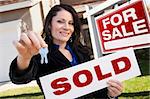 Happy Attractive Hispanic Woman Holding Sold Real Estate Sign and Keys in Front For Sale Real Estate Sign and House.