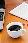 A cup of coffee with a laptop and a newspaper on a desk