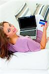 Smiling modern housewife sitting on sofa at home with laptop and credit card
