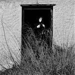 Masked figure by broken door of abandoned house obscured by overgrown plants.