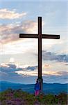 Small girl near wooden cross on summer heather flower hill top and evening country view behind