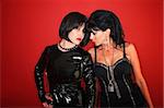 Two dominatrix women pose invitingly on a red background.