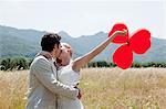 Newlyweds kissing in field with red heart shape balloons