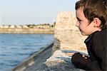 Little boy leaning against stone wall, looking at ocean view