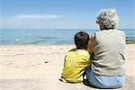 Grandmother and grandson sitting together on beach, looking at sea