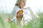 Barefoot young woman sitting in grass with dandelion flower between toes, cropped