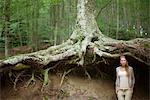 Woman standing at base of tree in woods, looking away in thought